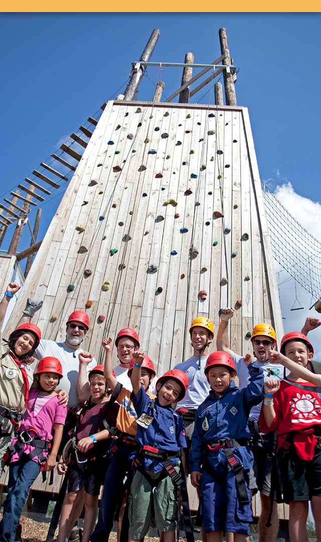 A group of Scouts wearing climbing gear cheering in front of the climbing tower